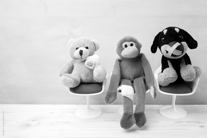 Three stuffed animals—a teddy bear with a bandaged arm, a monkey with a leg wrap, and a dog with a bandaged snout—sit on small white chairs, resembling patients in a waiting room. The scene conveys a playful, child-friendly healthcare environment.