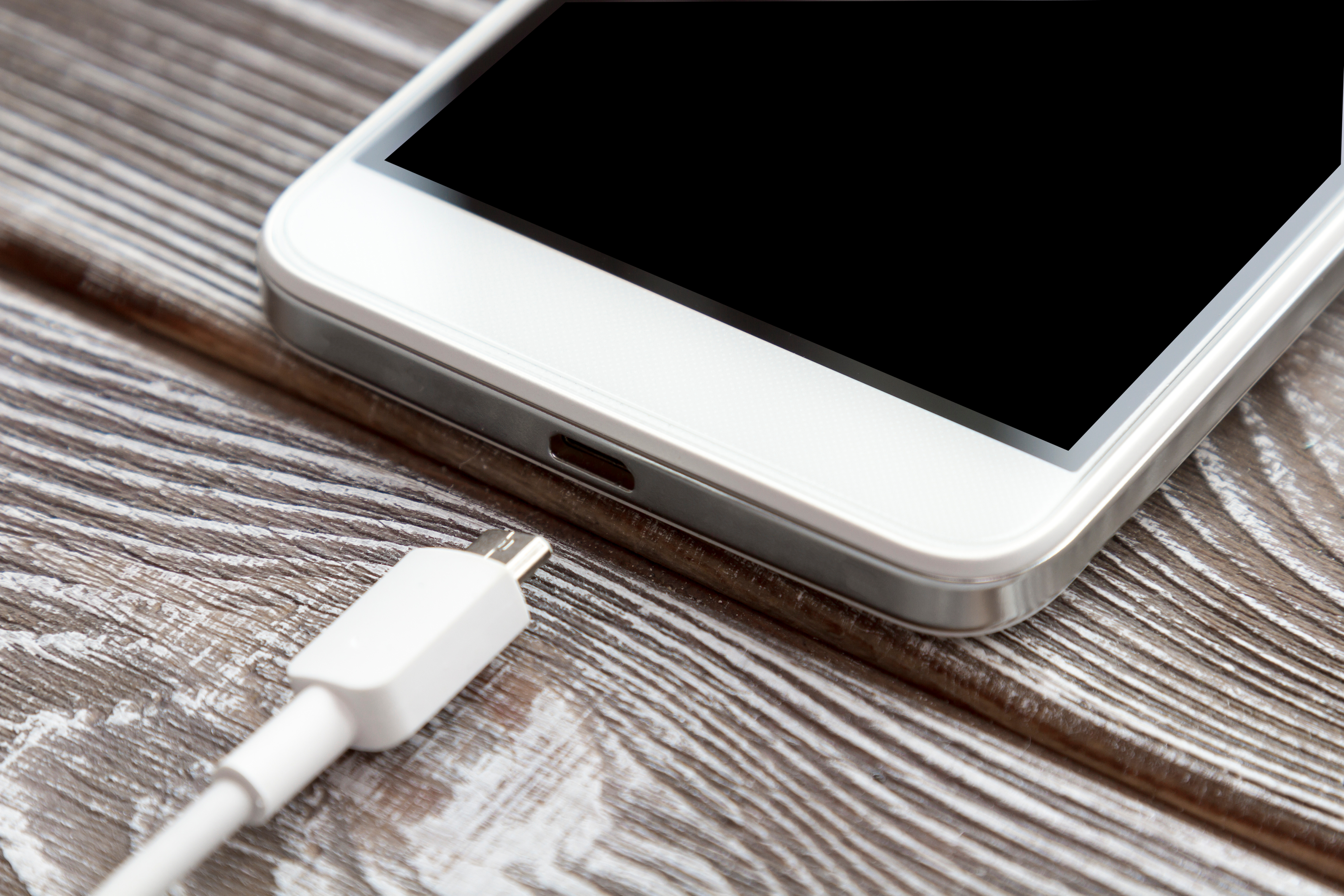 Close-up of a smartphone lying on a wooden surface with a USB charging cable positioned near the charging port, ready to be connected.