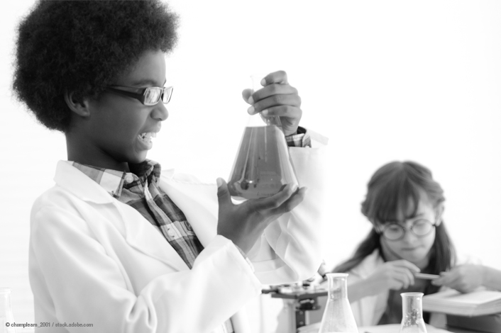 Person wearing a lab coat holding a conical flask with liquid in a science classroom, with another person working in the background near glassware and a notebook.