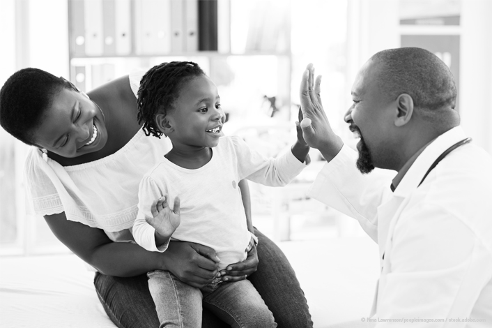 A smiling mother holds her young child on her lap during a medical visit. The child happily gives a high-five to a smiling healthcare provider, creating a warm and supportive atmosphere in the exam room.