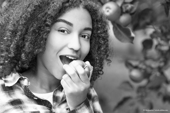 A teenage girl with curly hair smiles as she prepares to take a bite of an apple. She stands near an apple tree, wearing a plaid shirt