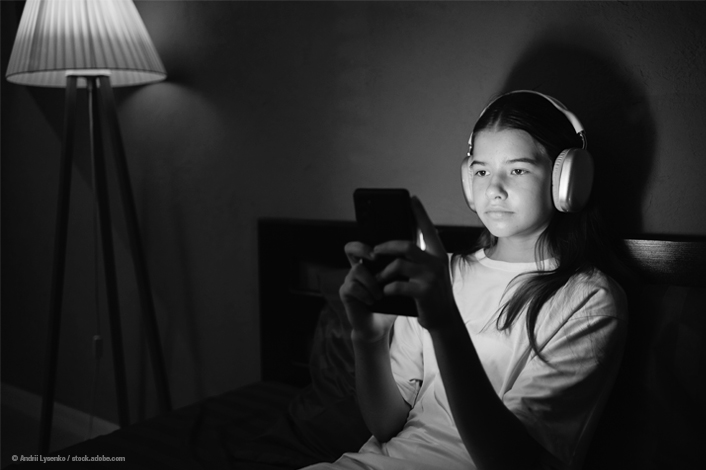 A young person sitting on a bed in a dimly lit room, wearing large headphones and holding a smartphone, illuminated by the phone’s light with a floor lamp glowing in the background.