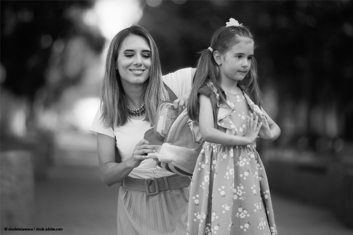 An adult helps a young girl adjust the straps on her backpack while they stand outdoors on a pathway. The girl is wearing a patterned dress and has a bow in her hair, while the adult is dressed in a skirt and short‑sleeved top. Trees and soft, out‑of‑focus lights line the background.