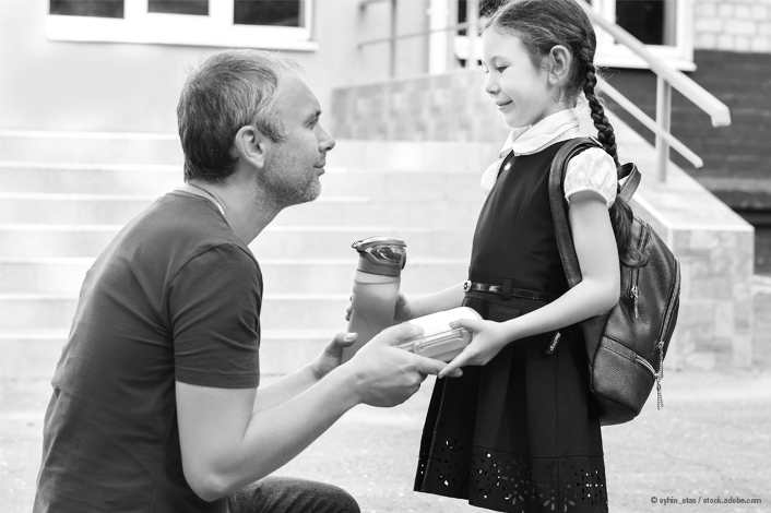 A man kneels in front of a young girl outside a building, handing her a lunch container while she holds a water bottle. The girl is wearing a dress and a backpack, and the two appear to be having a brief exchange before she heads off.