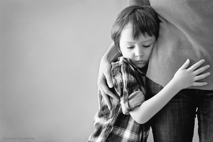 A young boy with a sad expression hugs an adult tightly around the waist. The adult gently holds the child, offering comfort and support.