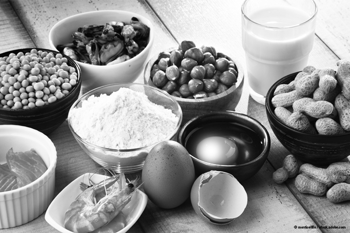A variety of common allergenic foods are arranged on a table, including eggs, milk, peanuts, soybeans, shellfish, flour, and nuts. The assortment represents ingredients often associated with food allergy testing.