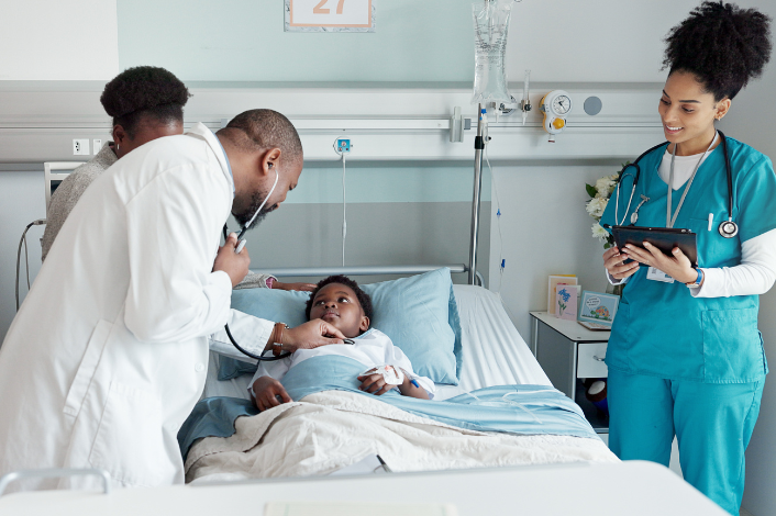 A medical team stands around a child lying in a hospital bed, with one clinician using a stethoscope to examine the patient while another reviews information on a tablet.