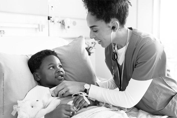 A healthcare professional uses a stethoscope to examine a child resting in a hospital bed. The child lies on pillows holding a stuffed toy, with medical tubing visible, in a bright clinical room suggesting a pediatric care setting.