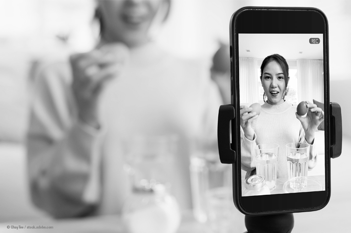 A smartphone mounted on a stand records a person seated at a table, holding two small round objects over several glasses of water. In the background, another person stands slightly out of focus, and the setting appears to be a bright indoor room, suggesting a video being recorded for social media or demonstration purposes.