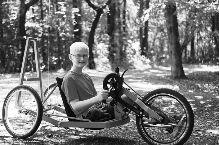 A smiling boy with light hair and glasses sits in a custom adaptive handcycle on a wooded trail. He wears a t-shirt and shorts, surrounded by trees and dappled sunlight