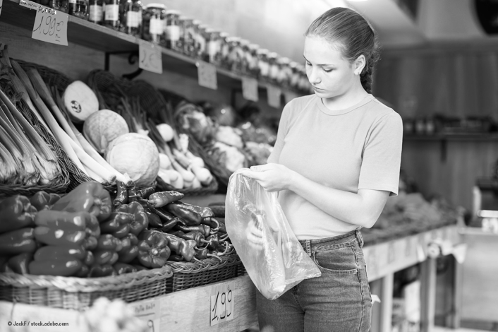A person stands in front of a market-style produce display, holding an empty plastic bag while selecting vegetables. The shelves are filled with assorted fresh items, including bell peppers, squash, leafy vegetables, and jars arranged in rows above the produce. The scene appears to be inside a small grocery or farm market, with wooden crates and price tags visible. The image is in black and white.