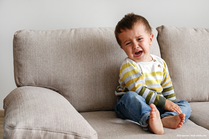 A small child sits on a light-colored couch with legs crossed and hands resting on the lap, wearing a striped long-sleeve shirt and jeans.
