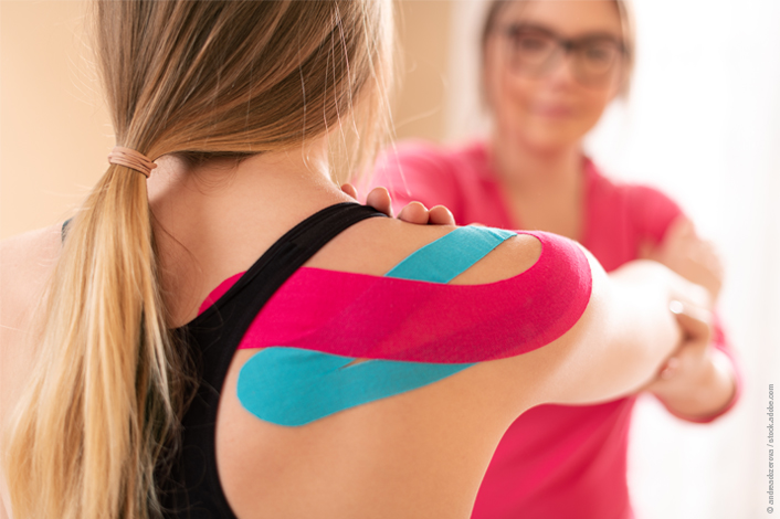 A young athlete’s shoulder is shown with pink and blue kinesiology tape applied. A clinician gently guides the arm during a physical therapy session