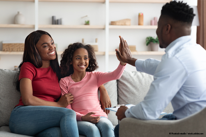 An adult and a child sit together on a couch during what appears to be a counseling or family meeting. A second adult sits across from them in a chair, raising a hand for a high‑five. Shelves with books and decorative items are visible in the background.