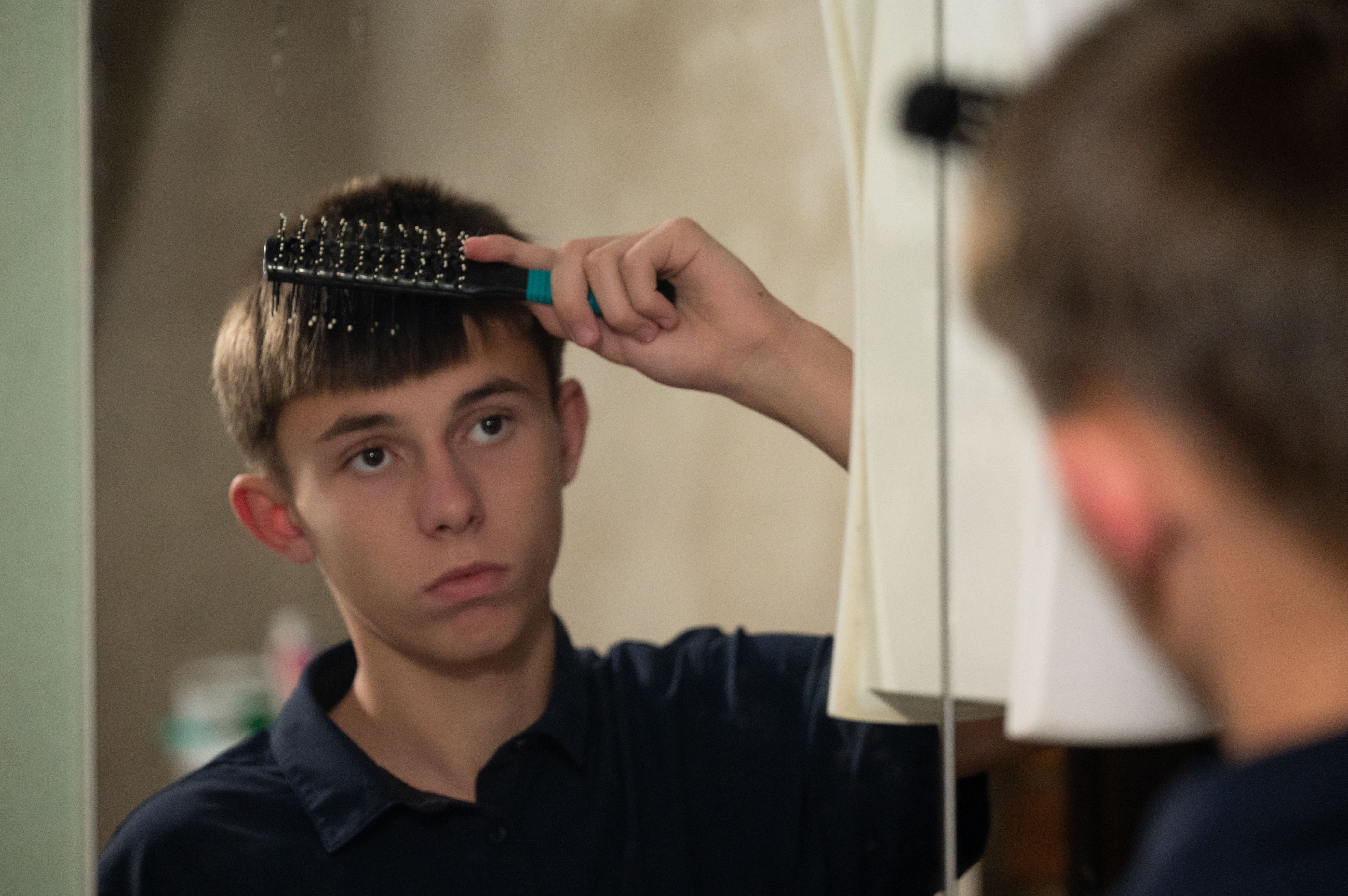 Person standing in front of a mirror brushing short hair with a black hairbrush. The brush is held near the top of the head, and the person’s reflection is visible in the mirror. The setting appears to be a bathroom or personal grooming space with neutral lighting.