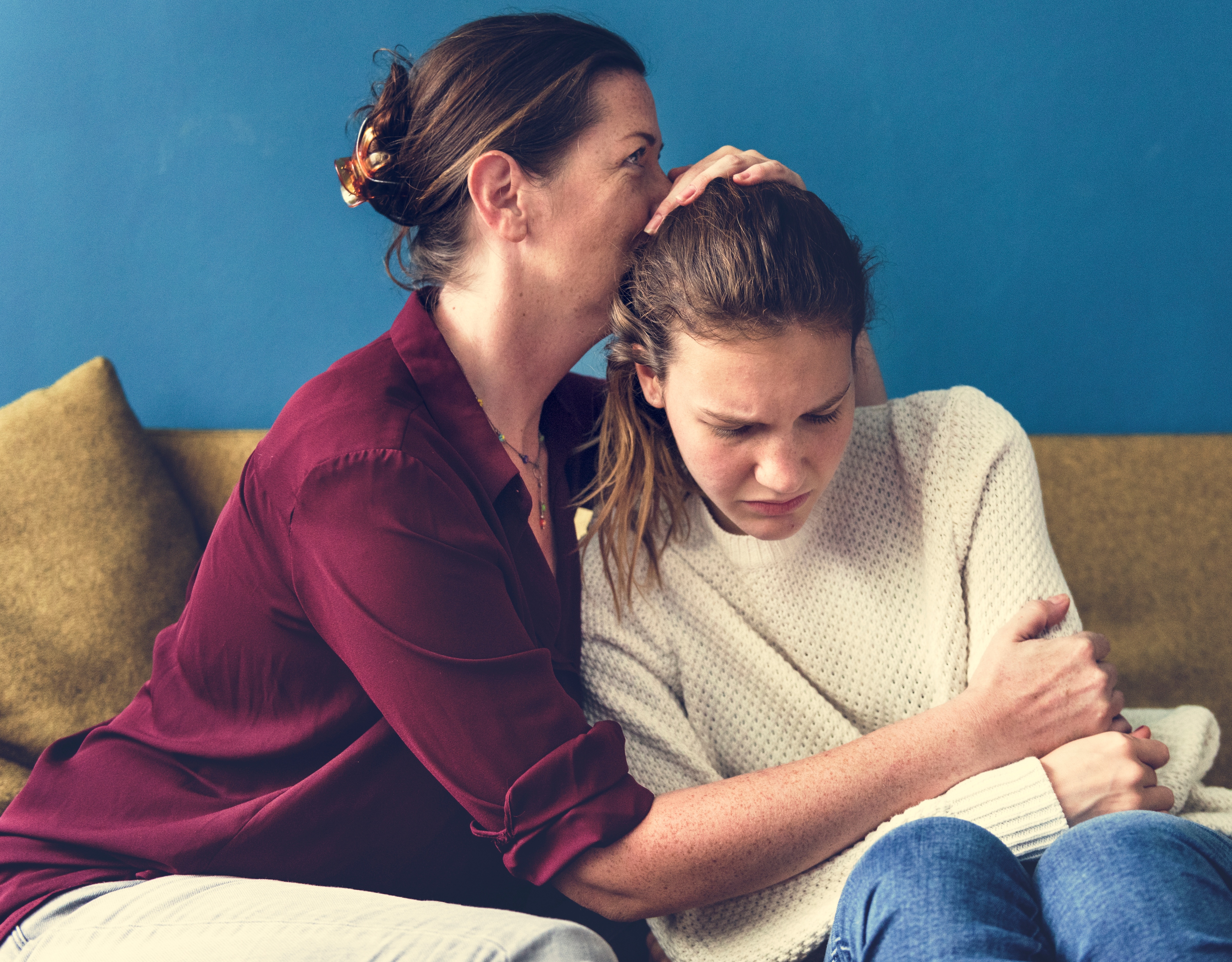 Two people sit together on a couch against a blue wall, with one person seated slightly behind and placing an arm around the shoulders of the other, resting a hand on the top of their head while the person in front sits leaning forward with arms crossed.