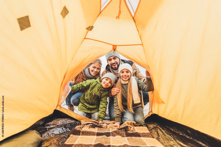 View from inside a bright orange tent looking outward at four people leaning into the entrance, with camping gear on the ground and an outdoor setting visible beyond the tent.