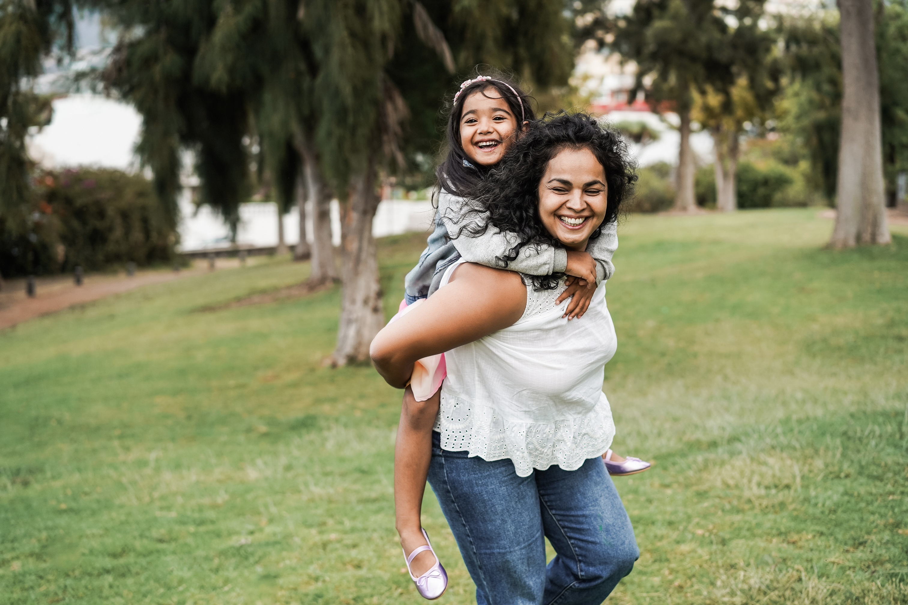 An adult carries a child on their back in a grassy park, with both facing the camera while trees and a walking path appear in the background.