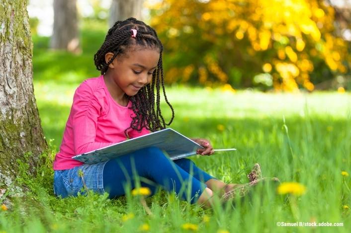 A young girl sits beneath a tree in a grassy park, smiling as she reads a large book. Sunlight filters through the trees, creating a peaceful outdoor setting that encourages a love of reading.