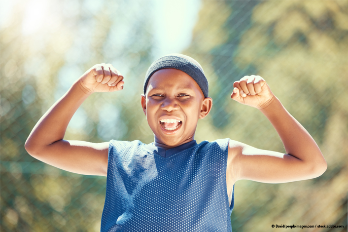 A young athlete smiles and flexes both arms outdoors while wearing a sleeveless sports jersey and head covering. The child’s confident pose and energetic expression emphasize strength, fitness, and injury prevention in youth sports.