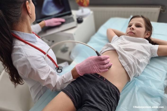 A healthcare provider performs an abdominal ultrasound on a child lying comfortably on an exam table. The child rests with hands behind their head while the clinician uses an ultrasound probe to examine the abdomen in a medical setting.