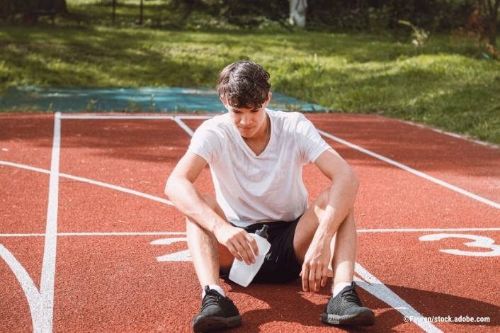A teenage athlete sits on a running track after exercise, holding a water bottle and looking down while resting. The image conveys fatigue, recovery, and sports-related health or injury management.