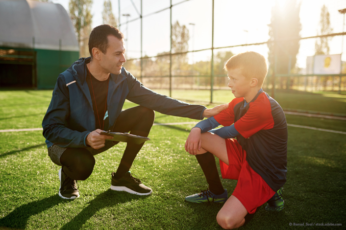 A coach talking to a middle school aged athlete while sitting on a field. 