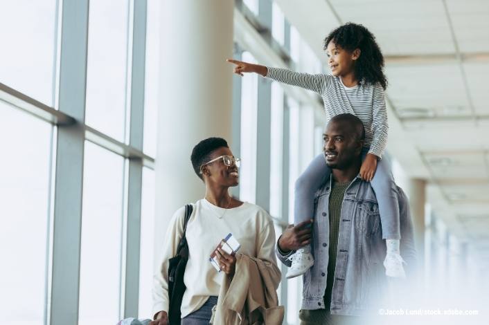 A family walks through an airport terminal with luggage. A young child rides on an adult’s shoulders and points ahead while another adult smiles beside them, conveying a positive and prepared travel experience.