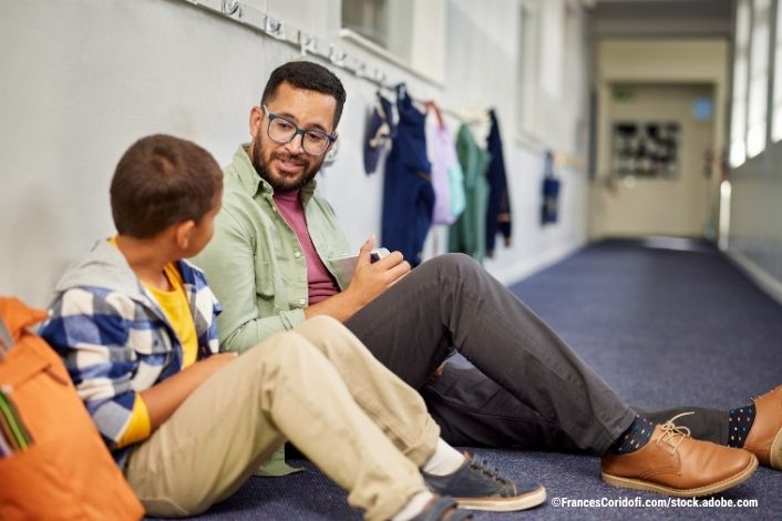 adult sitting on the floor at a school next to a young boy talking.