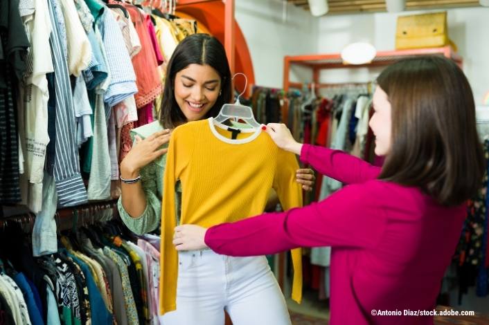 Teen girl wearing a purple sweater hold up a yellow sweater to another girl in a store