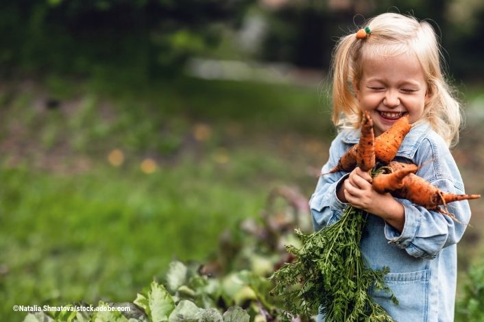 Toddler outside in a garden holding a bunch of carrots and smiling.