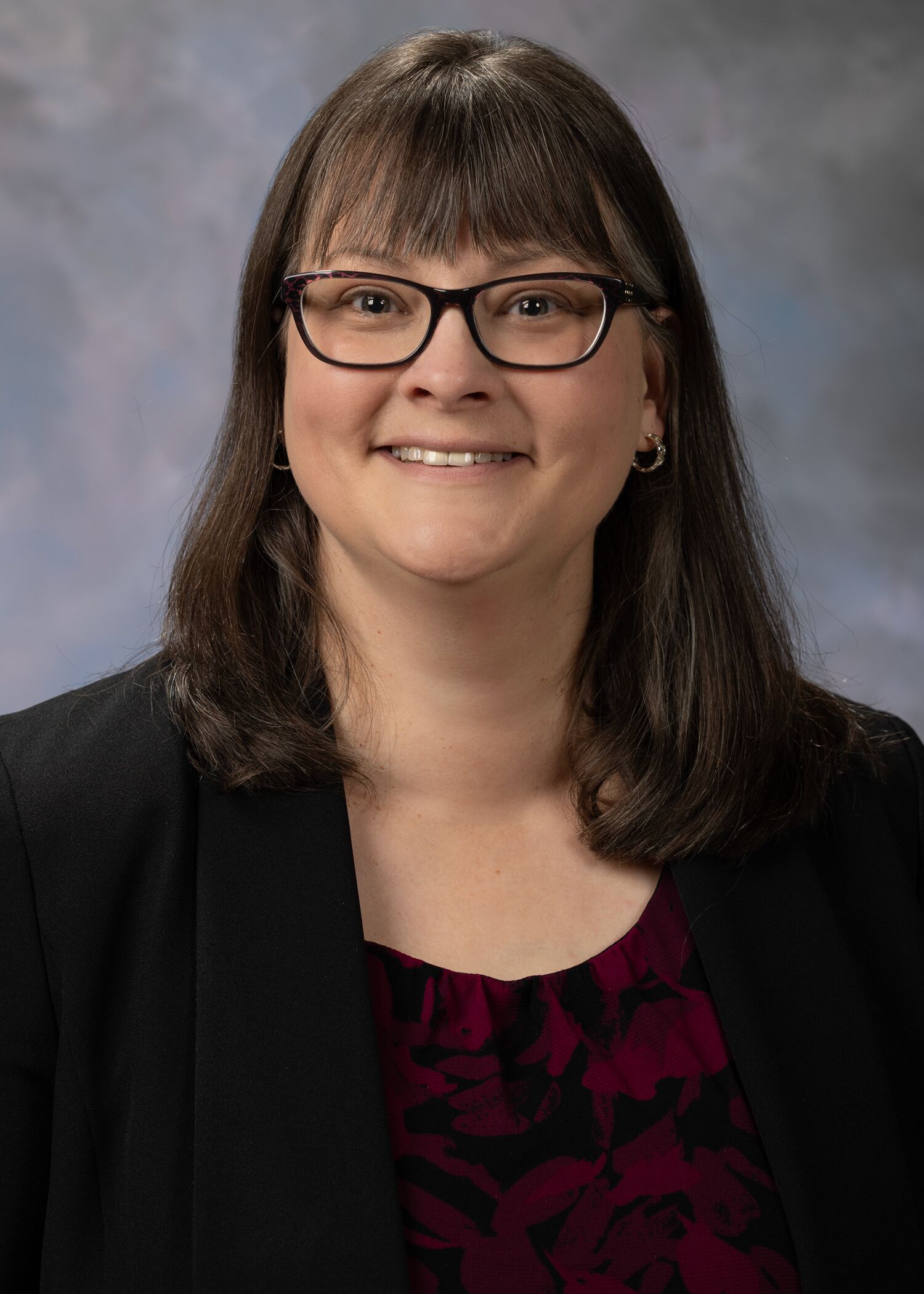 Portrait of Sarah Greenberg, woman with long brown hair, glasses, and a black blazer.