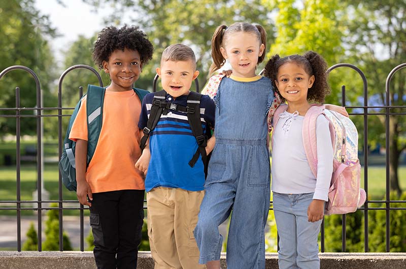 Smiling group of elementary school children wearing backpacks and standing together outdoors near a school, representing back-to-school readiness and childhood education.