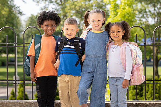 Four smiling children with backpacks standing together outdoors.