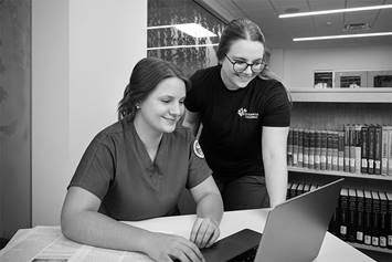 A nursing employee is seated in a library, with a laptop in front of her. A coworker stands behind her, smiling and looking at the laptop screen.