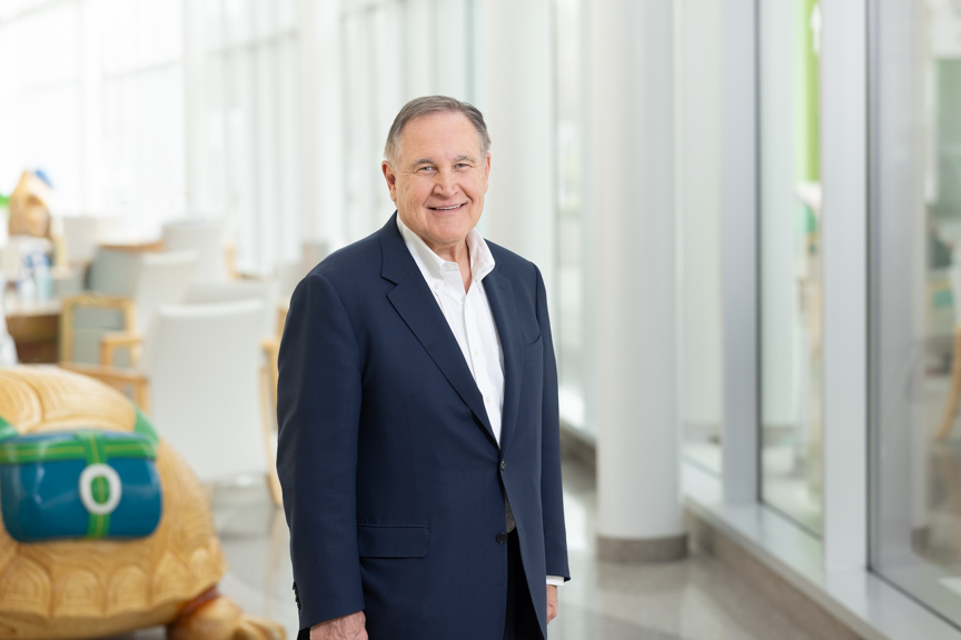 Tim Robinson smiles, wearing a dark suit and white shirt underneath. He's standing in a bright, light-filled space at the hospital.