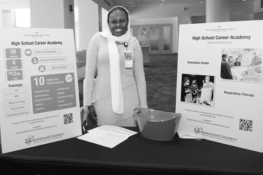 A female employee smiles, standing at a table. Informational posters about the High School Career Academy are displayed.
