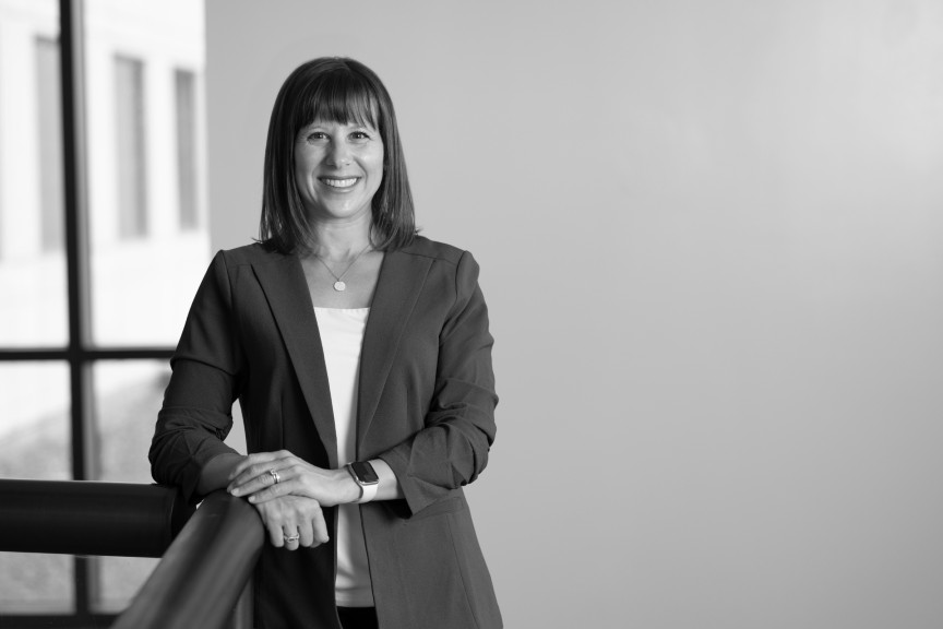 Christine is wearing a blazer. Her right elbow is resting on a railing.