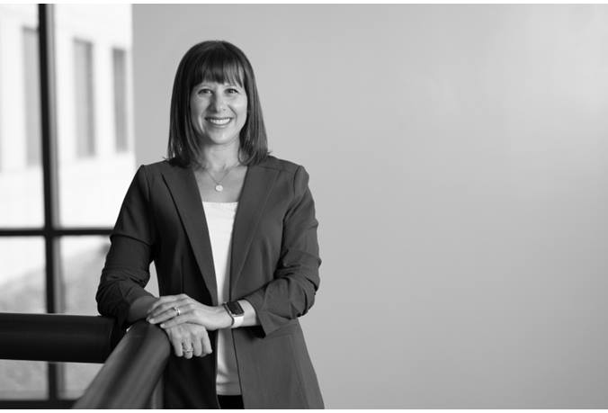 Christine is wearing a blazer. Her right elbow is resting on a railing.