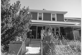 Alice, pictured wearing a mask, is standing on her front porch. The front of her home is pictured and there's a large tree in her front yard.