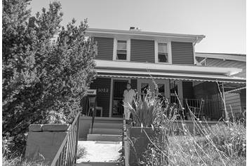 Alice, pictured wearing a mask, is standing on her front porch. The front of her home is pictured and there's a large tree in her front yard.