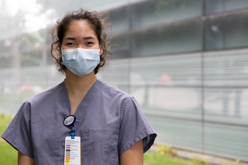 A teenage girl is standing outside, wearing a medical mask and scrubs, and smiling. 