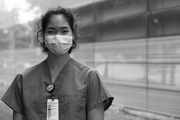 A teenage girl is standing outside, wearing a medical mask and scrubs, and smiling.