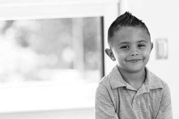 An elementary-aged boy is wearing a polo shirt, sitting in front of a window and smiling.