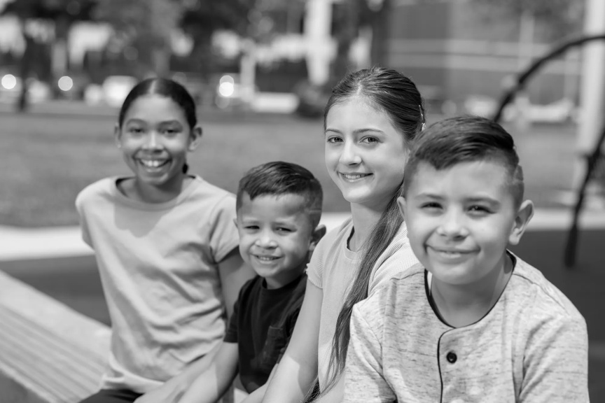 A group of preschool and elementary aged kids, two boys and two girls, are seated outside on a sunny, warm day. All are smiling.