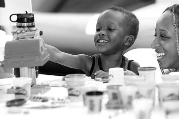 A young kid is seated at a table, playing and smiling. An adult is supervising and smiling.
