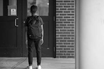 An older elementary/middle school-aged boy is wearing a backpack and walking toward a school's entrance. His back is facing the camera.