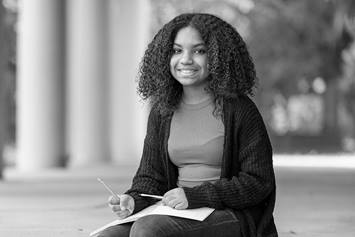 A teenage girl is wearing a cardigan and sitting on an outdoor stoop, writing in a notebook.