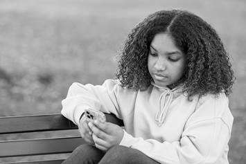 A teenage girl is relaxing while seated on a park bench, looking at her cell phone.