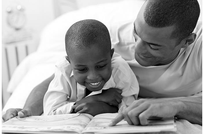 Dad is reading with his young son on the floor. He's pointing to something on the page and the son is smiling.